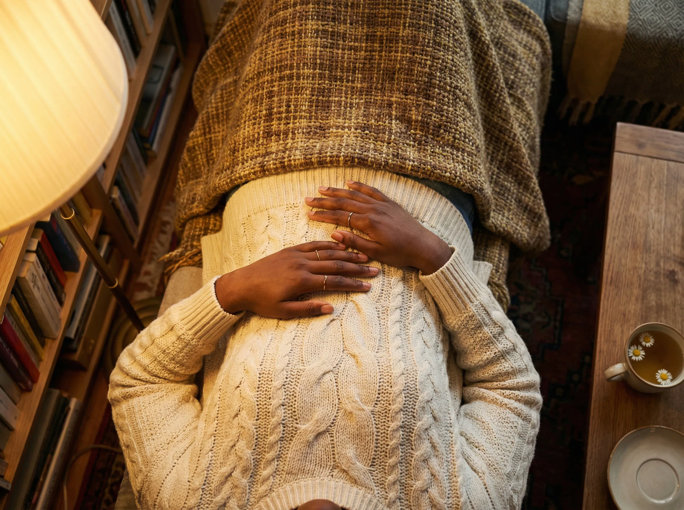 Ansicht von oben auf die Hände einer Frau, die während einer langsamen Atemübung auf ihrem Bauch ruhen, goldenes Lampenlicht über eine gestrickte Decke und Kamillentee in der Nähe