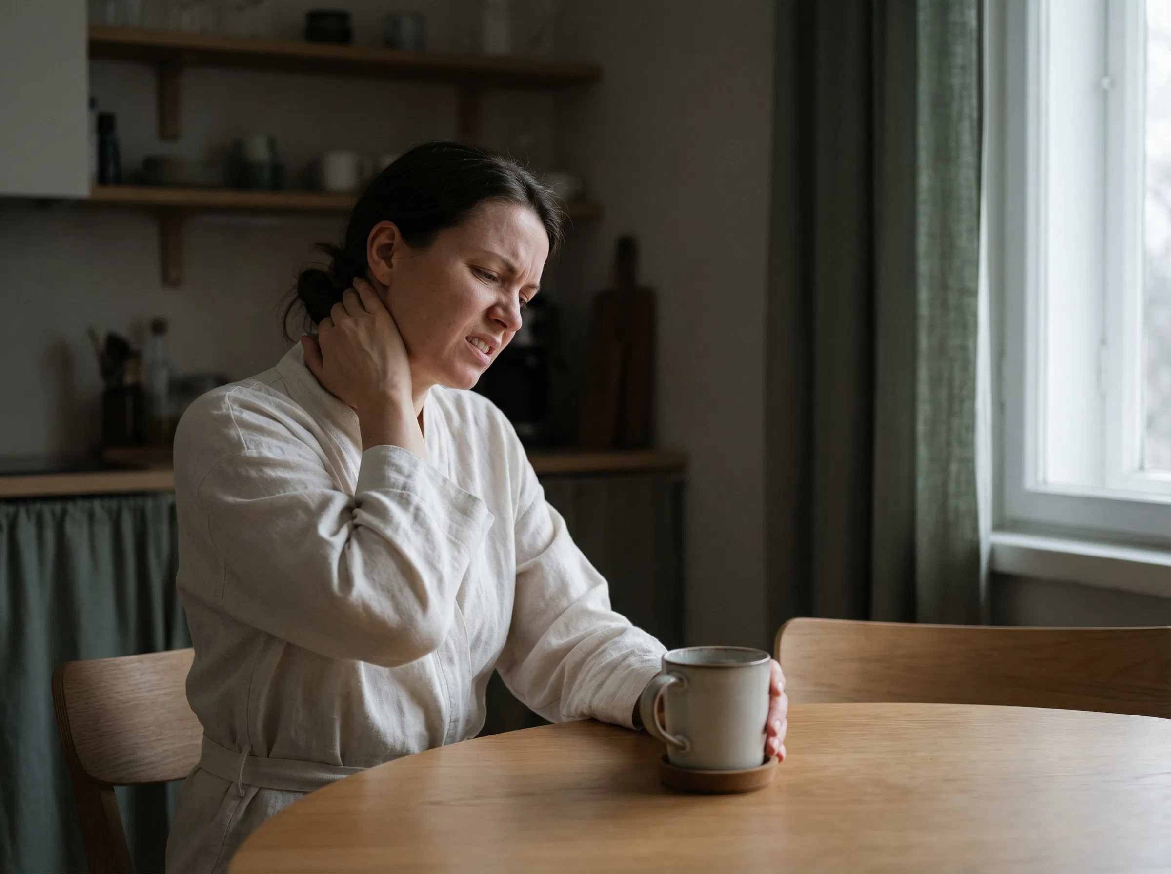 Mulher esfregando o pescoço com tensão na mesa da cozinha com um café frio intocado